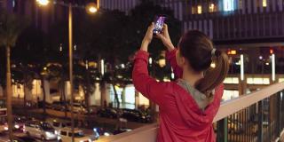 中国香港，一名年轻女子在晚上用手机拍摄建筑物。