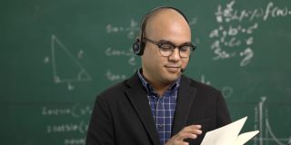 年轻的亚洲教师男子教授视频会议与学生。男性印度教师在课堂上通过在线课程培训数学。他戴着一副小小的谈话耳机。
