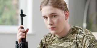 英俊的年轻军人肖像与步枪看着相机与严肃的面部表情。近距离特写的深思悲伤的白人士兵与武器在家里。