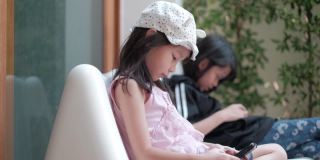 旁边拍摄的画面:两个泰国籍的亚洲女孩穿着休闲服装，专注于玩手机和平板电脑上的网络游戏。白天在花园区内。
