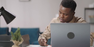 非裔美国学生在公寓里工作，在纸上写作，坐在桌子前用笔记本电脑，在线学习