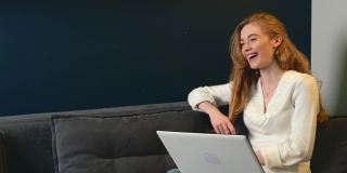 一名长着雀斑的红发女子躺在沙发上用笔记本电脑进行视频通话时面带微笑