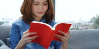 年轻的亚洲女人在温暖的家舒适的沙发上看书。放松女人拿着书在靠近窗户的沙发上看幸福。女人读书开书闲心。幸福女人生活在家里