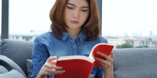年轻的亚洲女人在温暖的家舒适的沙发上看书。放松女人拿着书在靠近窗户的沙发上看幸福。女人读书开书闲心。幸福女人生活在家里