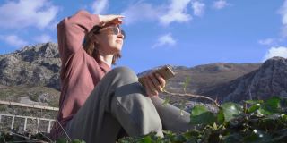 积极的女士戴着太阳镜凝视着远方，坐在古老的石头和绿色的植物，背靠山在蓝天下