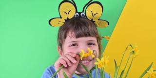 一个小女孩躲在绿色背景下的黄色水仙花后面。快乐的孩子戴着蜜蜂帽，拿着一盆花园花。春季是花园的播种季节。有选择性的重点