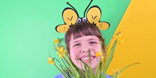 一个小女孩躲在绿色背景下的黄色水仙花后面。快乐的孩子戴着蜜蜂帽，拿着一盆花园花。春季是花园的播种季节。有选择性的重点