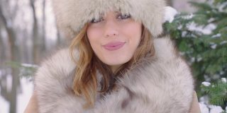 美丽的女人戴着皮帽看着相机在冬天的城市公园里白天在下雪的天气下飘落雪花，慢镜头