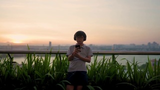 亚洲华人少年玩电话与耳机户外日落视频素材模板下载