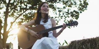 在清晨阳光的照射下，一名年轻美丽的亚洲女子穿着白衣在草堆上弹吉他。