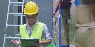 年轻微笑的黑人妇女工人在安全头盔和背心检查箱子在清洁工厂仓库使用板。女工程师准备发货和包装产品给客户。