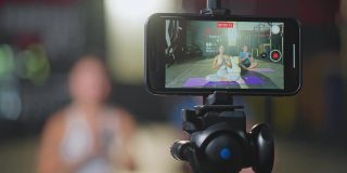 一名亚洲瑜伽女教师在健身房为一名白人男子授课。该女孩在网上直播自己的课程，并通过智能手机录制，以便在COVID - 19疫情期间健身房关闭时将学生隔离在家。