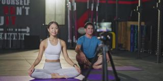 一名亚洲瑜伽女教师在健身房为一名白人男子授课。该女孩在网上直播自己的课程，并通过智能手机录制，以便在COVID - 19疫情期间健身房关闭时将学生隔离在家。
