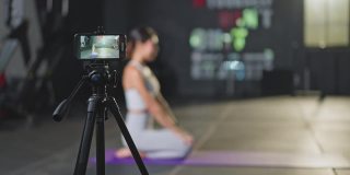 亚洲瑜伽女教师在健身房在线授课。该女孩在网上直播自己的课程，并通过智能手机录制，以便在COVID - 19疫情期间健身房关闭时将学生隔离在家。