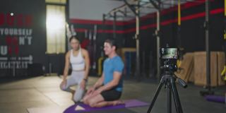 亚洲瑜伽女老师戴着面罩在健身房教一名白人男子，女孩在网上直播她的课，并通过智能手机录制，以便在COVID - 19疫情期间隔离学生在家。