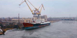 废弃的旧的生锈的船在船厂或码头与起重机，金属结构，码头在大型船舶建造设施的河流上融化的冰