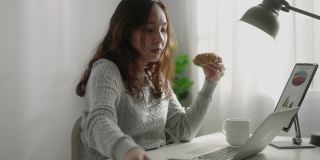 亚洲女商人一边用笔记本电脑工作一边吃甜甜圈