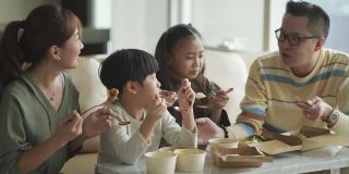 亚洲华人年轻家庭周末在客厅的电视前享受外卖食物