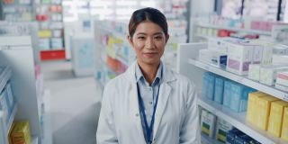 药店:美丽的亚洲药剂师的肖像穿着白大褂，看着相机和迷人的微笑，在她的摆满药包的货架后面。中等的特写镜头