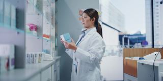 药店:美丽勤奋的亚洲药剂师的肖像穿着白大褂安排药包，药盒，维生素和补充药丸在柜台后面的架子上