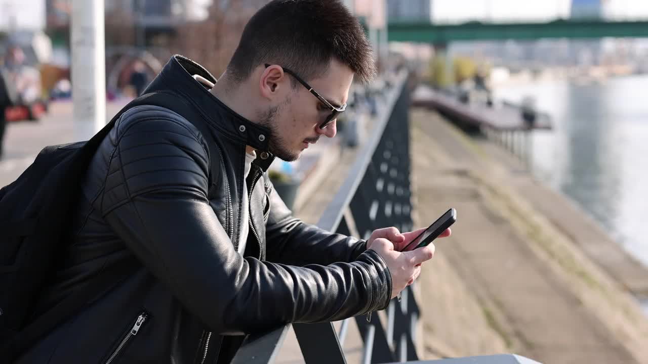 一个年轻人在城市的河边用手机发短信