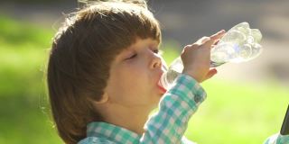 在夏天的公园里，快乐的小男孩喝着塑料瓶里的清水。孩子的肖像在户外饮水日落。关爱儿童保健，阳光热，特写
