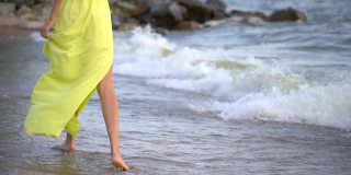 美丽的年轻女孩的腿穿着长黄色的衣服赤脚走在沙滩上的沙子靠近风微风海浪海岸快乐的假期旅游旅游度假女人旅行者享受自然