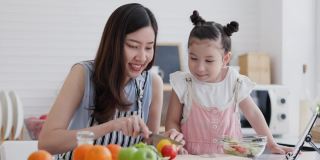 家庭单身母亲和女儿享受和快乐的烹饪在干净的厨房。家长帮助孩子制作水果和蔬菜沙拉的视频教程从平板电脑。通过视频电话学习自制烹饪。