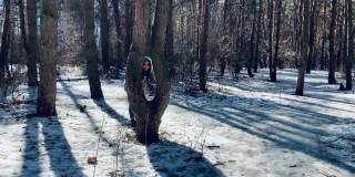 穿着灰色大衣的年轻女子在森林里的树林里向外张望。在早春的一个阳光明媚的日子里，漂亮的雌性躲在树后