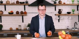 戴着眼镜、穿着西装、心情愉快的年轻商人坐在厨房里摆弄着两个橙子。慢镜头，从工作中休息