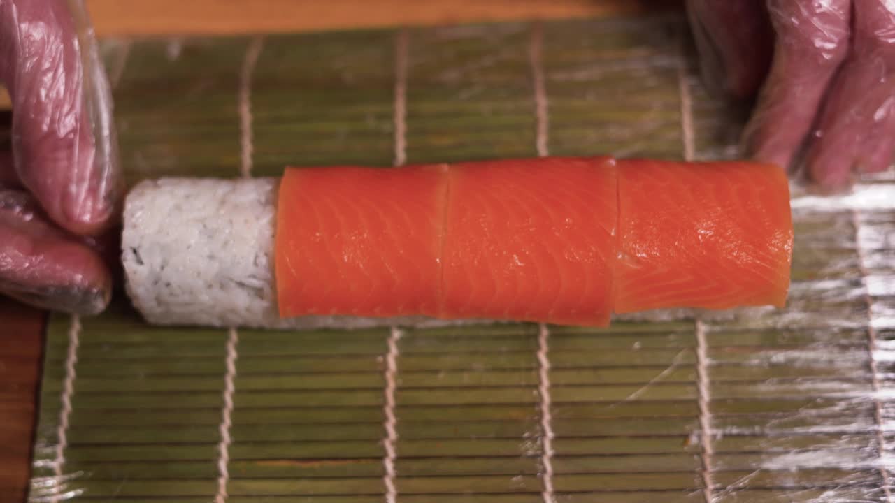 一位专业厨师将切成薄片的三文鱼放到餐桌上的寿司卷上。在一家日本餐厅，男厨师正在准备一个三文鱼寿司卷。