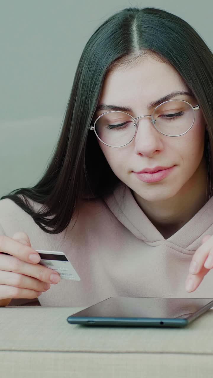 一名年轻的千禧一代白种女孩戴着眼镜，在疫情期间使用数字平板电脑和信用卡在家进行网上购物。