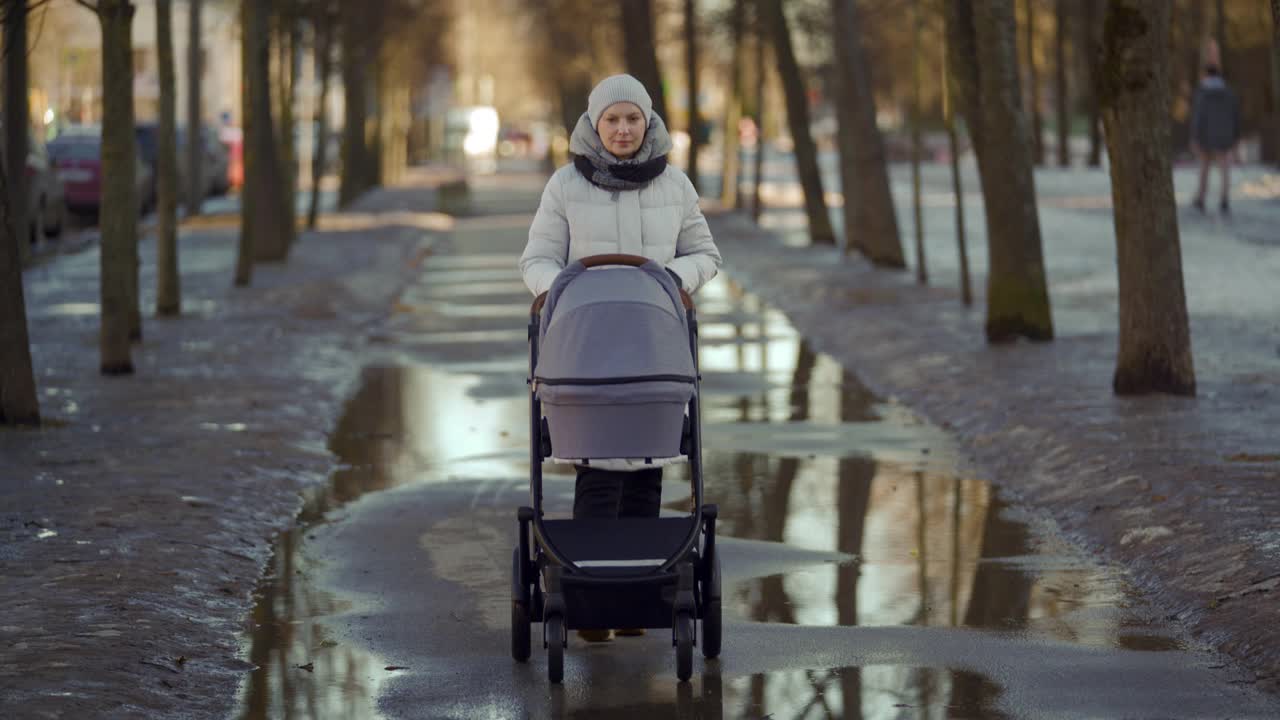 母亲推着婴儿车走在街上，妇女推着婴儿车走在镇上的小路上，冬天或早春季节