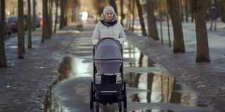 母亲推着婴儿车走在街上，妇女推着婴儿车走在镇上的小路上，冬天或早春季节