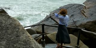 一个戴着棕色帽子的女孩站在多石的河岸上看着海浪