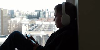 十几岁的男孩戴着白色耳机，穿着黑色兜帽夹克，坐在窗台上，享受着从他的手机听音乐。保持社交距离，在家娱乐