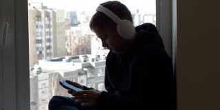 十几岁的男孩戴着白色耳机，穿着黑色兜帽夹克，坐在窗台上，享受着从他的手机听音乐。保持社交距离，在家娱乐