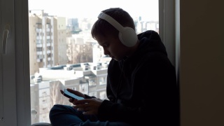 十几岁的男孩戴着白色耳机，穿着黑色兜帽夹克，坐在窗台上，享受着从他的手机听音乐。保持社交距离，在家娱乐视频素材模板下载