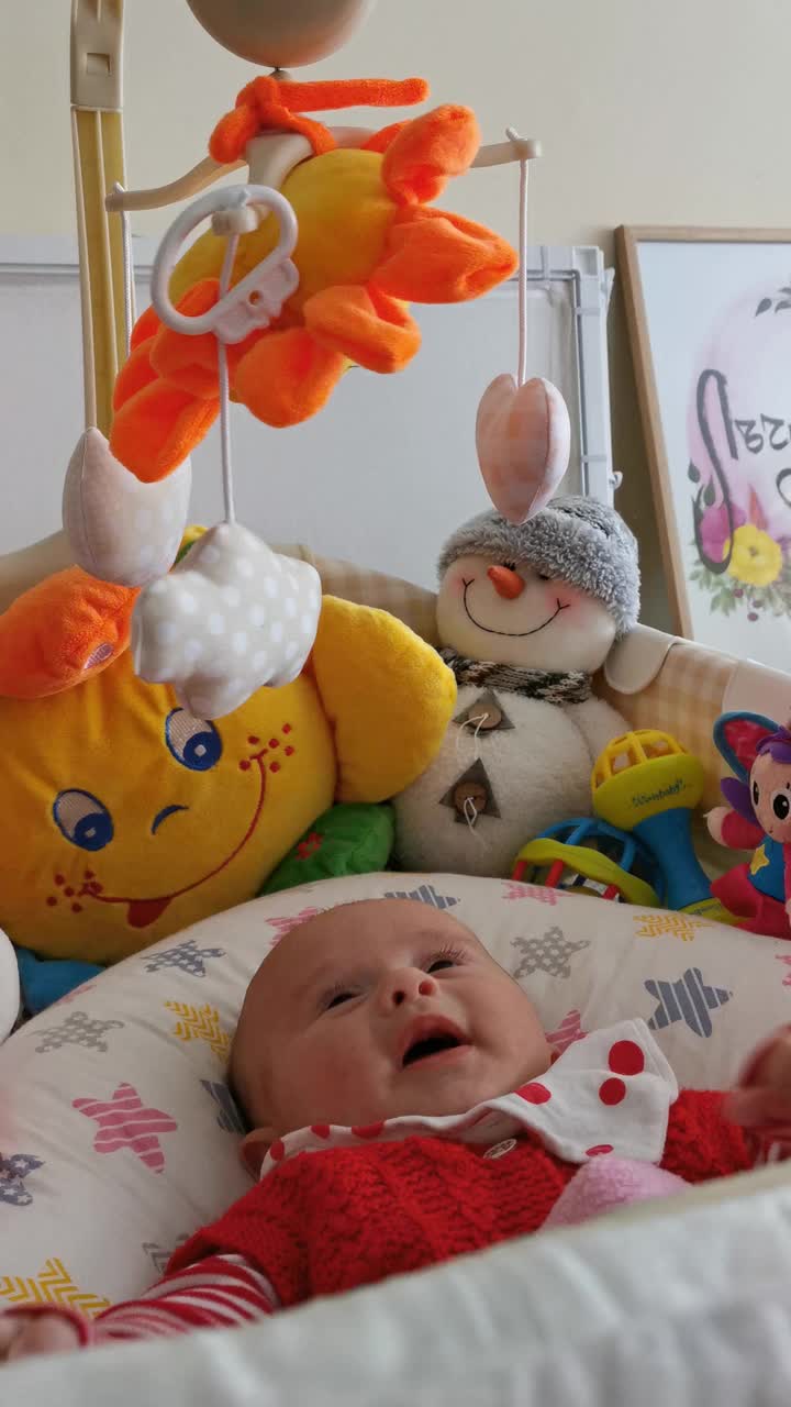 微笑的婴儿看着她的玩具，笑着看着玩具旋转。三个月大的女婴在床上玩耍。COVID-19大流行期间的新生儿。
