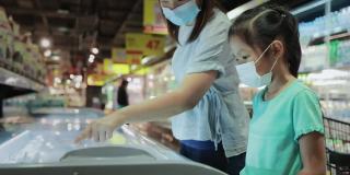 一名亚洲女童和她的母亲戴着防护口罩，以预防冠状病毒大流行，新的正常要求是一起选择超市冷藏室的冷冻食品。