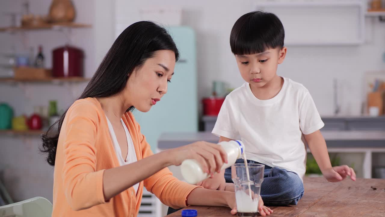 迷人的年轻母亲微笑着为她的儿子在家里吃早餐倒牛奶。