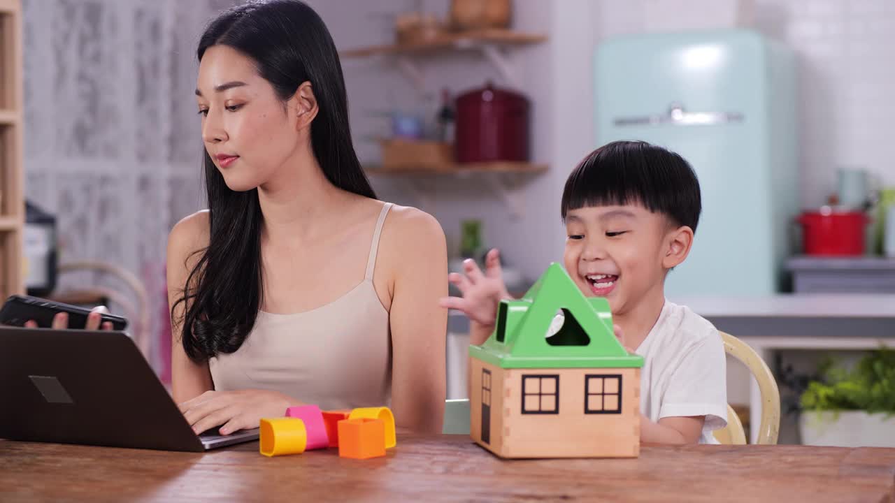 忙碌的母亲一心多用，亚洲年轻的母亲正在用手机和笔记本电脑工作，小儿子坐在她旁边，在家里的餐桌上玩小塑料房子玩具。