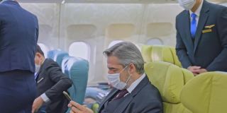 新冠肺炎疫情期间戴口罩乘坐飞机预防感染的白种商务旅客。乘务员通知乘客关闭电子设备。