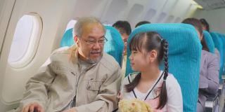 亚洲爷爷和女儿坐在飞机上，乘飞机旅行度假。女孩抱着小熊，在飞机飞行时和爸爸玩手机游戏。航空运输的概念