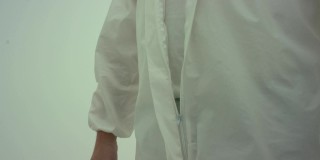 戴着深色圆框眼镜的男人拉上防护服的拉链。特写镜头跟随双手，拍摄在白色背景上