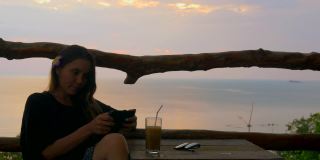 日落时分，一个美丽的女孩在一家俯瞰大海的咖啡馆里，正在用智能手机
