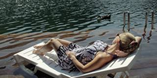 一个女人躺在太阳床上，戴着太阳镜，披着波西米亚风格的丝绸披肩。女孩休息在一个洪水木头水下码头。鸭子和天鹅在附近游泳。