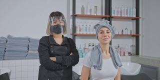 肖像亚洲职业女性造型师双手交叉，戴口罩和面罩以防止冠状病毒感染，站在年轻的白人顾客坐在沙龙洗床