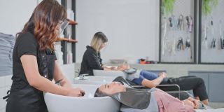 亚洲年轻美丽的女人顾客躺在沙龙洗床头发洗发廊由发型师。大流行期间，发型师佩戴面罩，防止冠状病毒感染