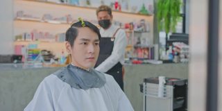 亚洲专业男性发型师在美发厅为年轻顾客梳理和使用剪刀理发。大流行期间，发型师佩戴防护口罩，防止冠状病毒感染。